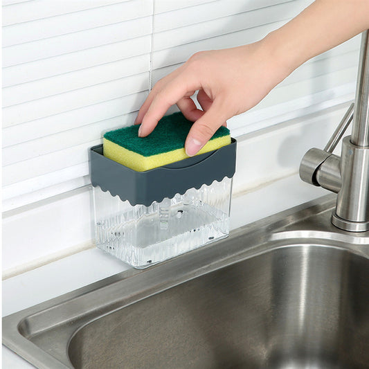 The image shows a clear, ribbed 2-in-1 soap dispenser and sponge caddy positioned next to a kitchen sink. A person's hand is pressing a yellow and green sponge into the gray top plate of the dispenser, which is designed to dispense soap onto the sponge when pressed.