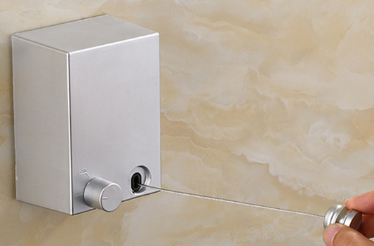 The image shows a sleek, silver retractable clothesline device mounted to a marble-textured wall. A metallic knob is being pulled, extending a thin line from the box, illustrating the product's function and compact design.