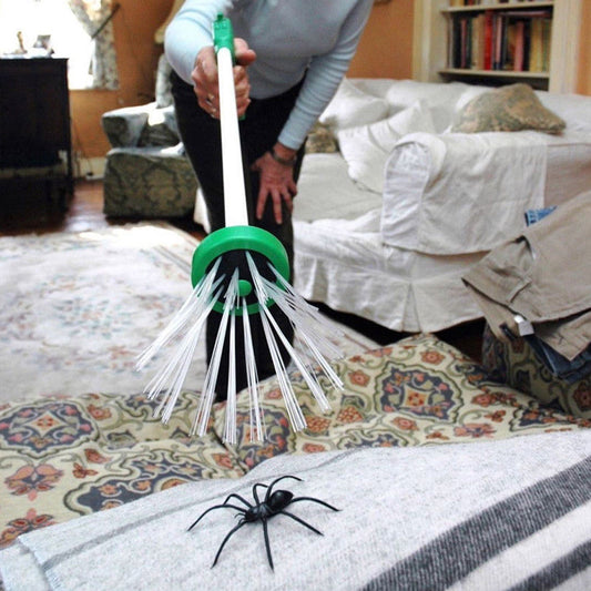 The image shows a person using a long-handled insect catcher to trap a large, black spider on a striped gray and white blanket. The catcher has a round, green top with white bristles that gently encompass the spider without harm. The background includes a cozy room with a bookshelf and a white sofa.