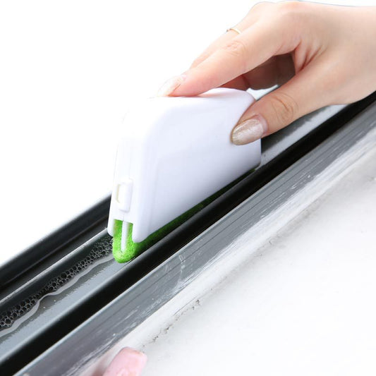 The image shows a close-up of a white handheld cleaning brush with a green scouring pad, being used to clean the narrow groove of a window frame. The window frame is white and the brush fits snugly into the groove, indicating its design for such small spaces.