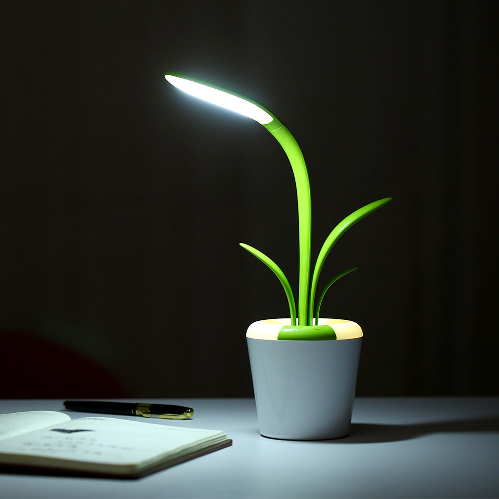 image displays a single green Clivia LED Table Lamp. The lamp, resembling a stylized plant, has a white, pot-like base and two green, leaf-shaped elements that extend upward. One leaf is illuminated and curved like a flexible stem, providing a bright white light over an open notebook on a dark background.