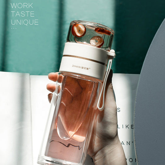 The image shows a sleek, transparent glass water bottle with a double-wall design, filled with a brown liquid, possibly tea, with two visible ice cubes. The bottle has a white and beige silicone band around its center for grip and a matching portable rope handle. It's set against a blurred background with soft teal tones, possibly suggesting a tranquil, modern setting