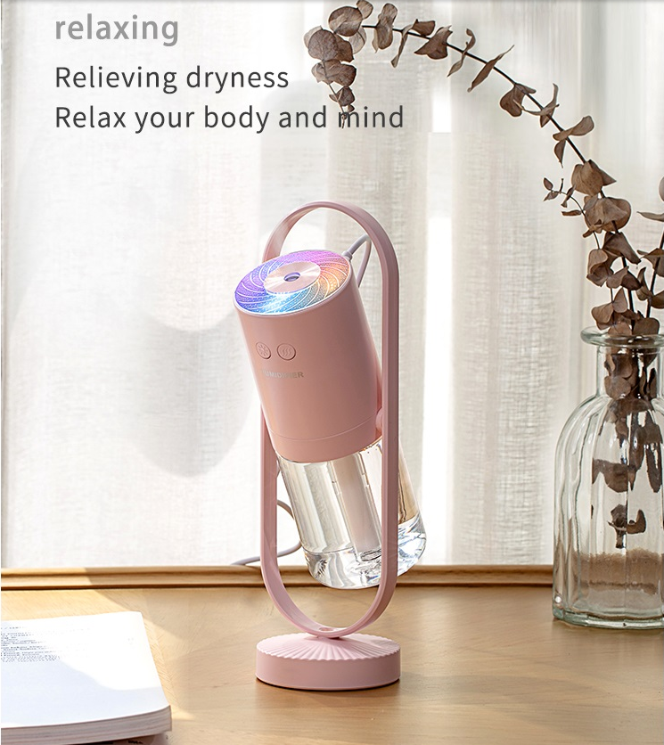 The humidifier, in a pastel pink shade, is displayed on a wooden table emitting a colorful glow from the top, suggesting a relaxing ambiance.