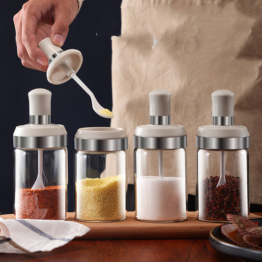 The image displays a set of four glass seasoning jars with stainless steel caps, aligned neatly on a wooden surface. Each jar contains a different seasoning, visible through the clear glass, with one jar being used by a hand with a spoon to extract some powdered seasoning.