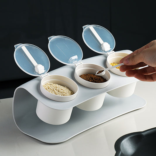 The image displays a Creative Seasoning Box Combination Set placed on a counter, with three compartments each housing different spices, topped with clear, flip-open lids and a spoon for dispensing.