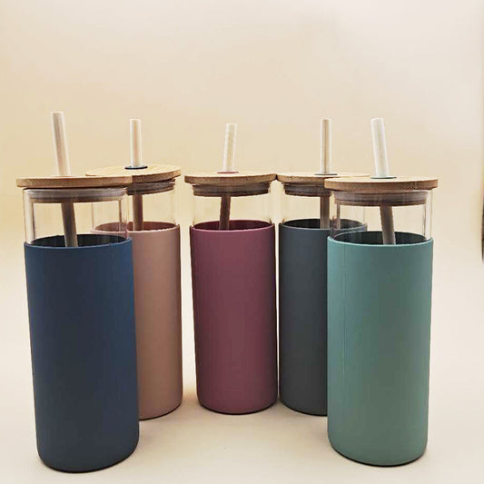 Four cylindrical glass cups with matte finishes in navy, maroon, teal, and grey, each with a wooden lid and a clear glass straw inserted through the center.