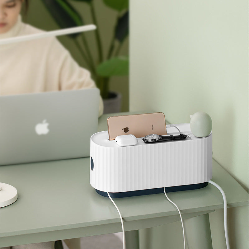 The Cloud Power Storage Box in white is displayed on a green table, with a person placing a beige phone on top. It showcases the box's spacious top, ideal for holding phones and small items, and emphasizes its role as a cable organizer.