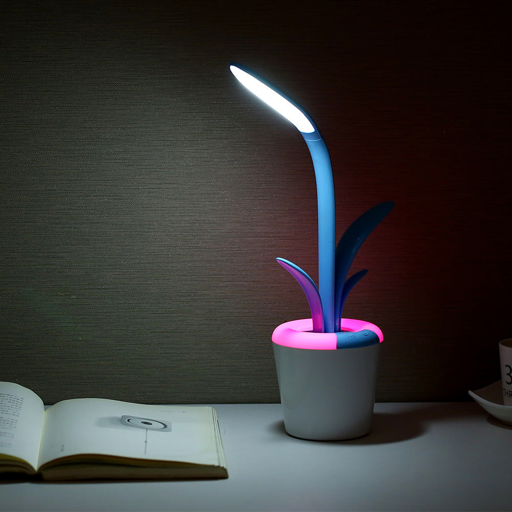  image features a blue Clivia LED Table Lamp with a pink lighted base, adding a colorful glow to the scene. The lamp has a similar design to the first, with a white base and plant-like appearance, casting a bright white light onto an open book placed on a table with a textured brown background.