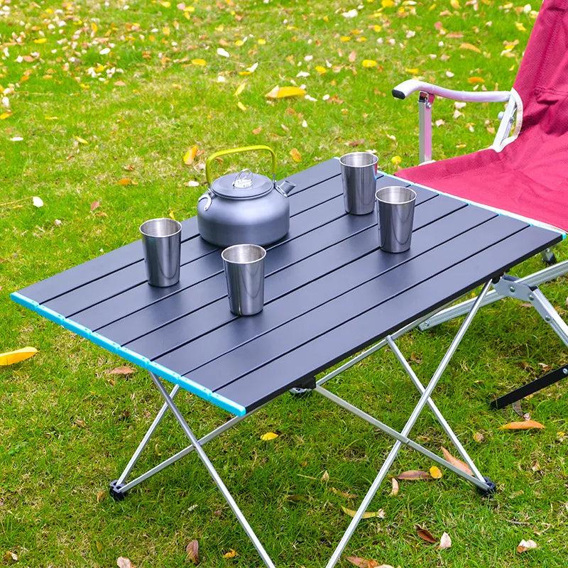 blue folding camping table set in a grassy outdoor area: "This lightweight camping table in vibrant blue is ideal for outdoor dining, showcasing a sturdy aluminum frame and foldable design for easy transport to picnics or garden parties