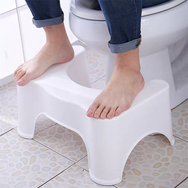 The image displays a simple yet functional white plastic bathroom stool placed in front of a toilet. It has a distinctive U-shape design that curves around the base of the toilet, with two levels for foot placement. The stool appears sturdy and is designed to assist in achieving a natural squat position. It is used by a person wearing blue jeans rolled up to the ankles, indicating how the stool might be used during toilet use to support the feet for better posture and comfort.