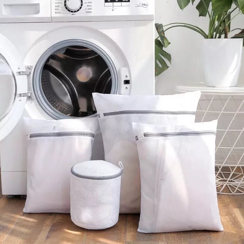 The image displays a variety of mesh laundry bags in front of a washing machine. There are three rectangular bags in different sizes with secure zippers on top and a cylindrical bag with a lid, all designed for washing delicates. The bags appear to be made of fine white mesh material, allowing for visibility and breathability of the contents.