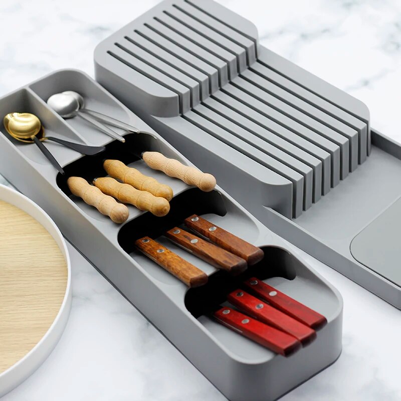 The image shows a plastic cutlery storage tray in grey, with compartments for knives, spoons, and forks. Knives with wooden and red handles rest in a slotted holder, while gold and silver spoons lie in a separate compartment. The organizer is on a marble surface, suggesting a kitchen setting.