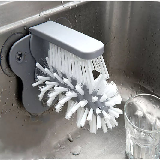 The image depicts a gray-colored, wall-mounted cup scrubbing device in action, with water flowing over its bristles, which are actively cleaning a transparent glass placed upside down on the brush. The scrubber is attached to the inside of a stainless steel sink, demonstrating its functionality and ease of use in a kitchen setting.