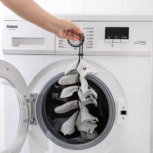 The image shows a hand holding a black adjustable clothesline with several pairs of grey socks hanging from it. The clothesline is hooked onto the open door of a white front-loading washing machine, showcasing the product's use in a laundry setting.