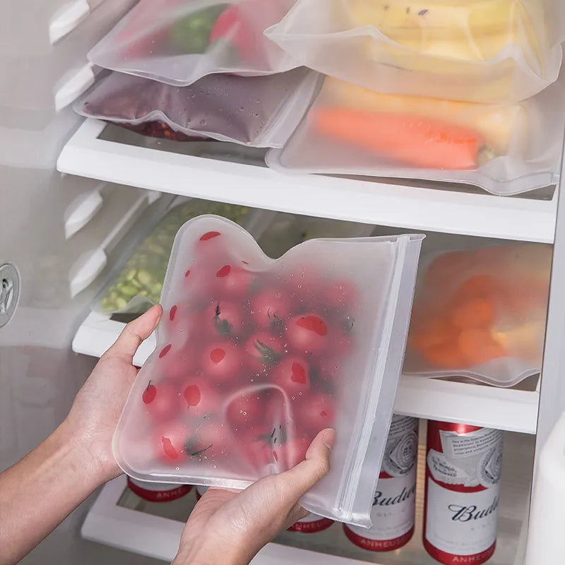 Hands holding a translucent silicone storage bag filled with red berries, with a refrigerator shelf background.