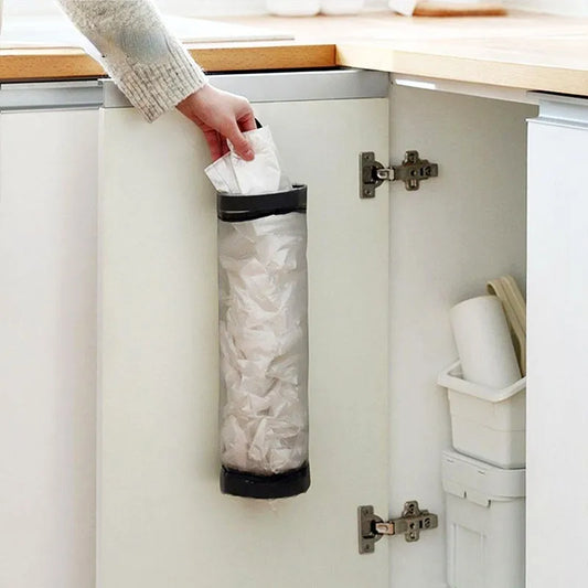 The image displays a wall-mounted plastic bag holder affixed to a light-colored cabinet door. It appears cylindrical in shape, with a black band around the top and bottom, filled with white plastic bags. A person's hand is seen depositing a bag into the holder, indicating its ease of use.