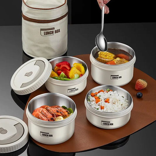The image shows a collection of stainless steel insulated lunch boxes with multiple compartments, each containing different food items such as fruits, vegetables, rice, and shrimp. The containers are open and placed on a dark surface, with a beige carrying bag labeled "LUNCH BOX" partially visible in the background.