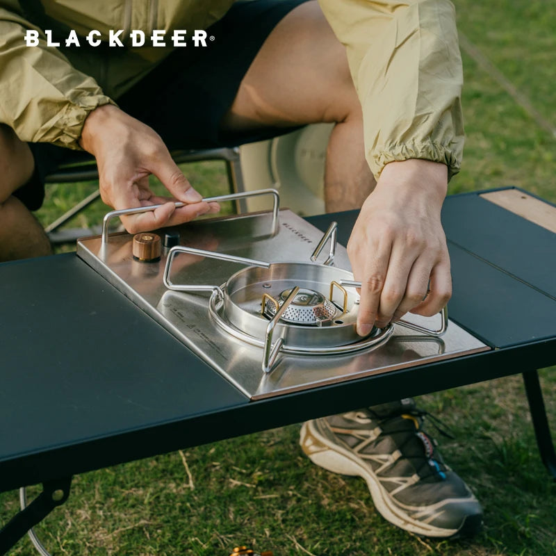 A user is adjusting a portable stove on the BLACKDEER Traveler Modular Combination Desk. This foldable picnic table is perfect for campsite cooking or outdoor gatherings.