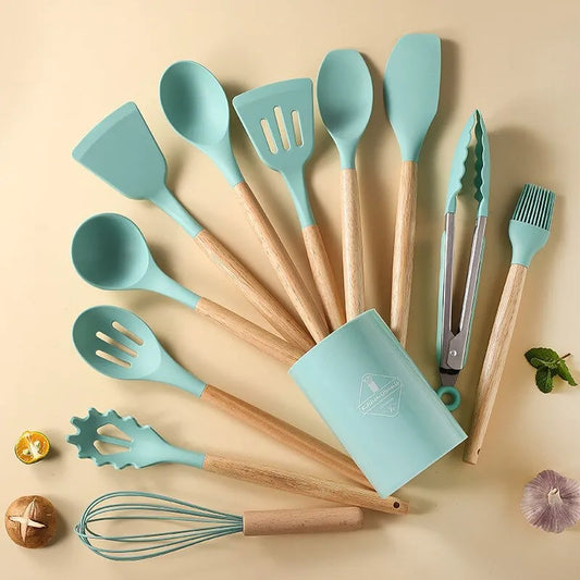 This image displays a set of pastel teal silicone kitchen utensils with light wooden handles. The set includes a variety of tools such as spatulas, spoons, a ladle, a whisk, and tongs, along with a container for storage. The utensils are arranged neatly on a pale background, accompanied by a few scattered cooking ingredients.