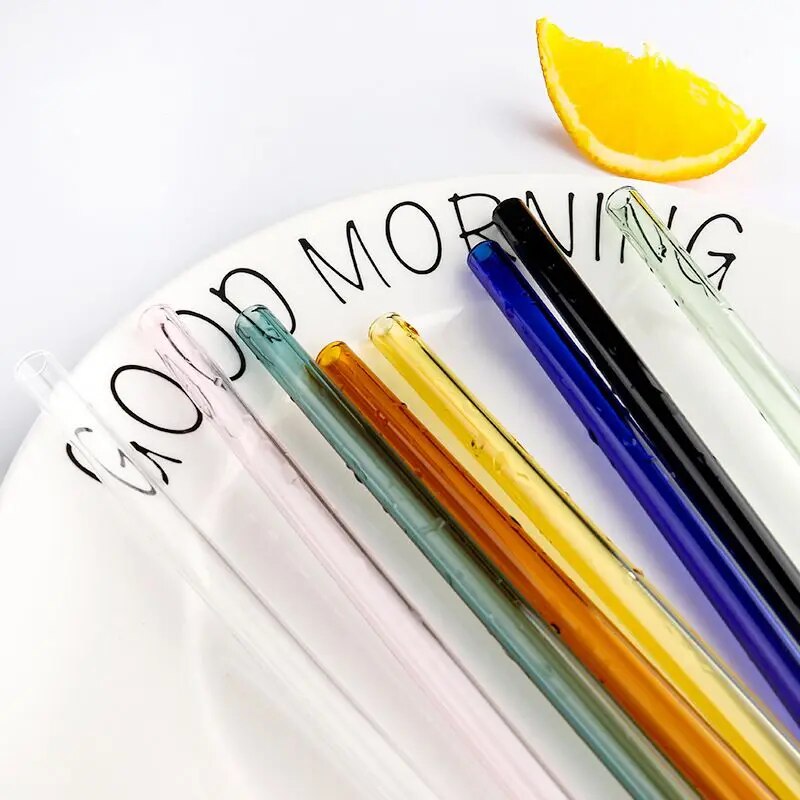 Assorted glass straws with transparent, blue, green, yellow, and red colors laid out on a white surface with text "GOOD MORNING" next to a lemon slice