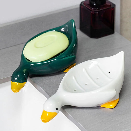 The image shows two ceramic soap dishes, each shaped like a duck. One dish is white with yellow details on the beak and feet, while the other is a dark green with the same yellow accents. Both dishes feature a slight incline and grooves to allow water to drain, keeping the soap dry.