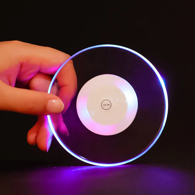 This image depicts a hand holding a round, acrylic coaster that emits a luminous glow around the edges. The coaster features a LED light that shifts colors, currently set to a soft purple hue, enhancing the ambiance of any setting where a drink might be placed.