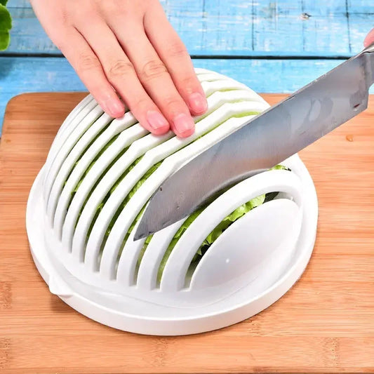 The image depicts a hand using a knife to slice through a white, dome-shaped salad cutter bowl filled with leafy greens. The bowl has slits to guide the knife, ensuring even cuts. This kitchen tool rests on a wooden cutting board set against a blue-painted background.