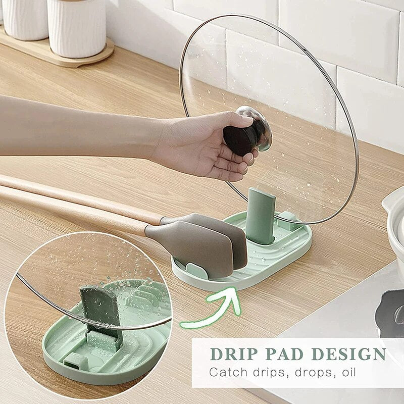 The image shows a hand lifting a transparent pot lid from a sage green silicone pot lid holder. The holder, which also supports a spatula, has a grooved design to catch drips, drops, and oil, preventing mess on the countertop.