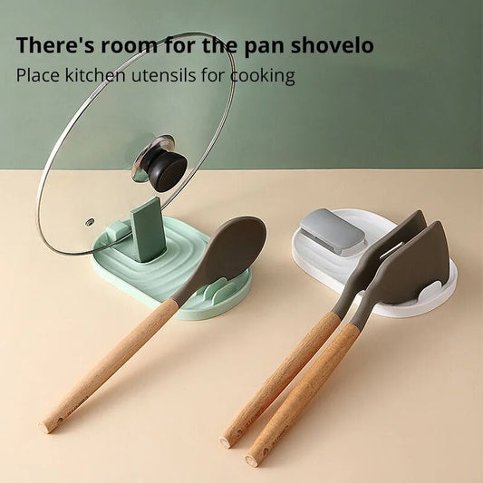 The image displays two silicone pot lid and utensil holders, one in sage green and the other in grey, against a dual-tone background. A wooden spoon rests on the green holder, and a spatula on the grey, illustrating the product's use in a kitchen setting.