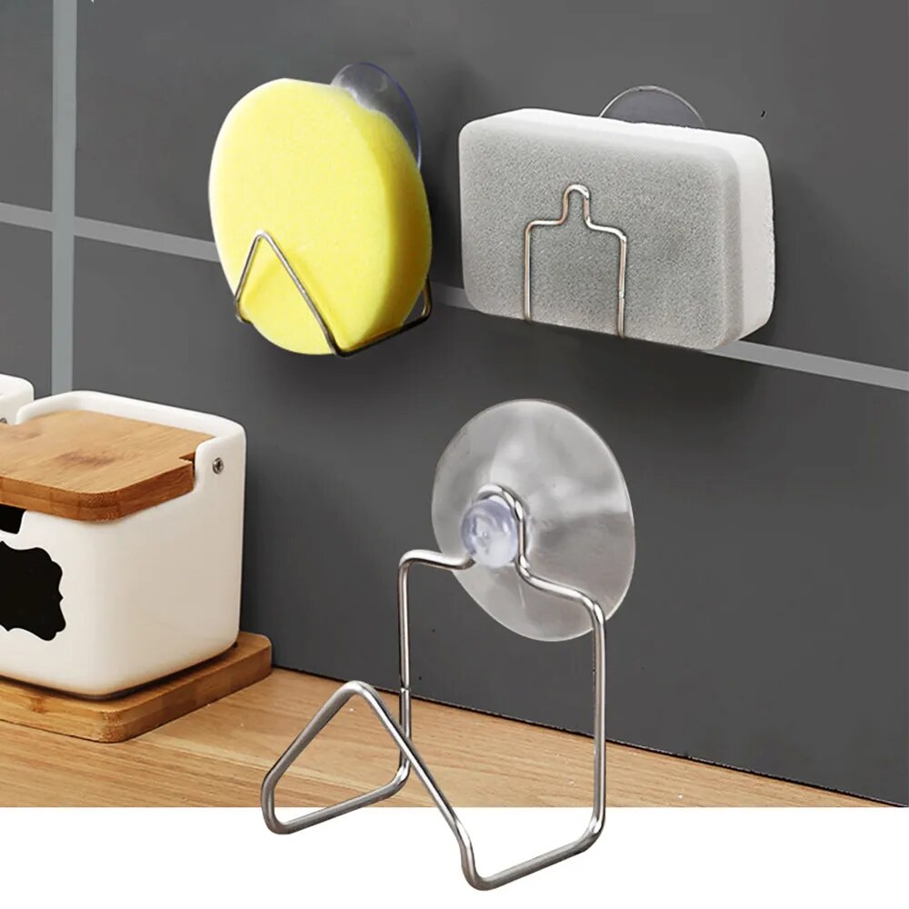 The image shows three suction cup-based drain racks mounted on a grey wall. Two racks are holding sponges: one is yellow and the other is grey. The third rack, with no sponge, is made of stainless steel and has a minimalist design. The kitchen environment suggests utility and organization.