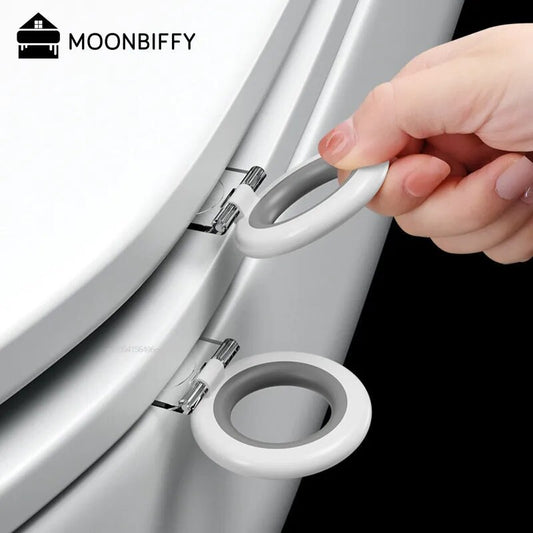 The image shows a close-up of a white toilet seat with a silver-toned attachment mechanism. A person's hand is lifting a white, oval-shaped toilet seat lifter that's affixed to the edge of the seat for hygienic handling. The device appears sleek and is designed to minimize contact with the toilet for cleanliness.