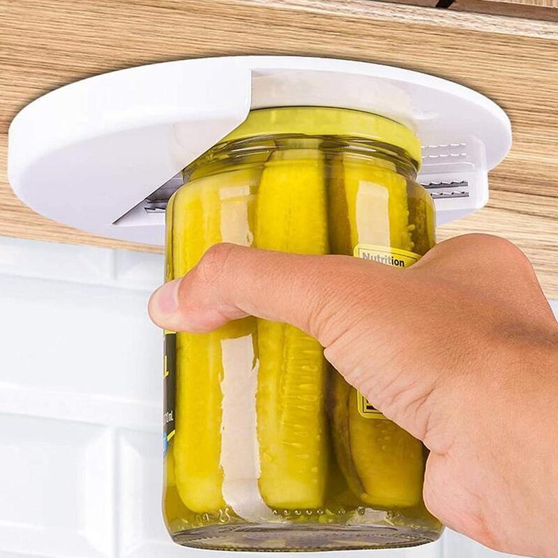 Another close-up of a hand holding a jar of pickles, the jar’s lid being gripped by the white opener mounted under a cabinet.